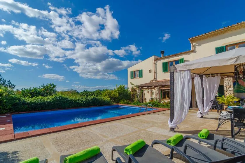 Un patio soleado junto a una piscina, con asientos y una pérgola con cortinas.
