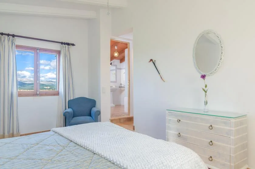 Un dormitorio con una cama, un sillón azul y una cómoda junto a la ventana.