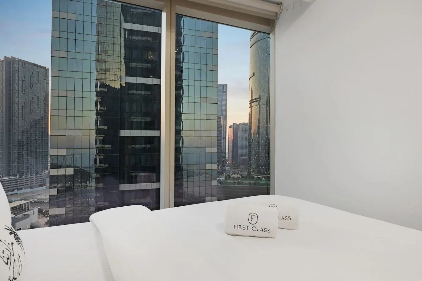 Dormitorio con una cama junto a grandes ventanas con vistas a edificios de la ciudad.