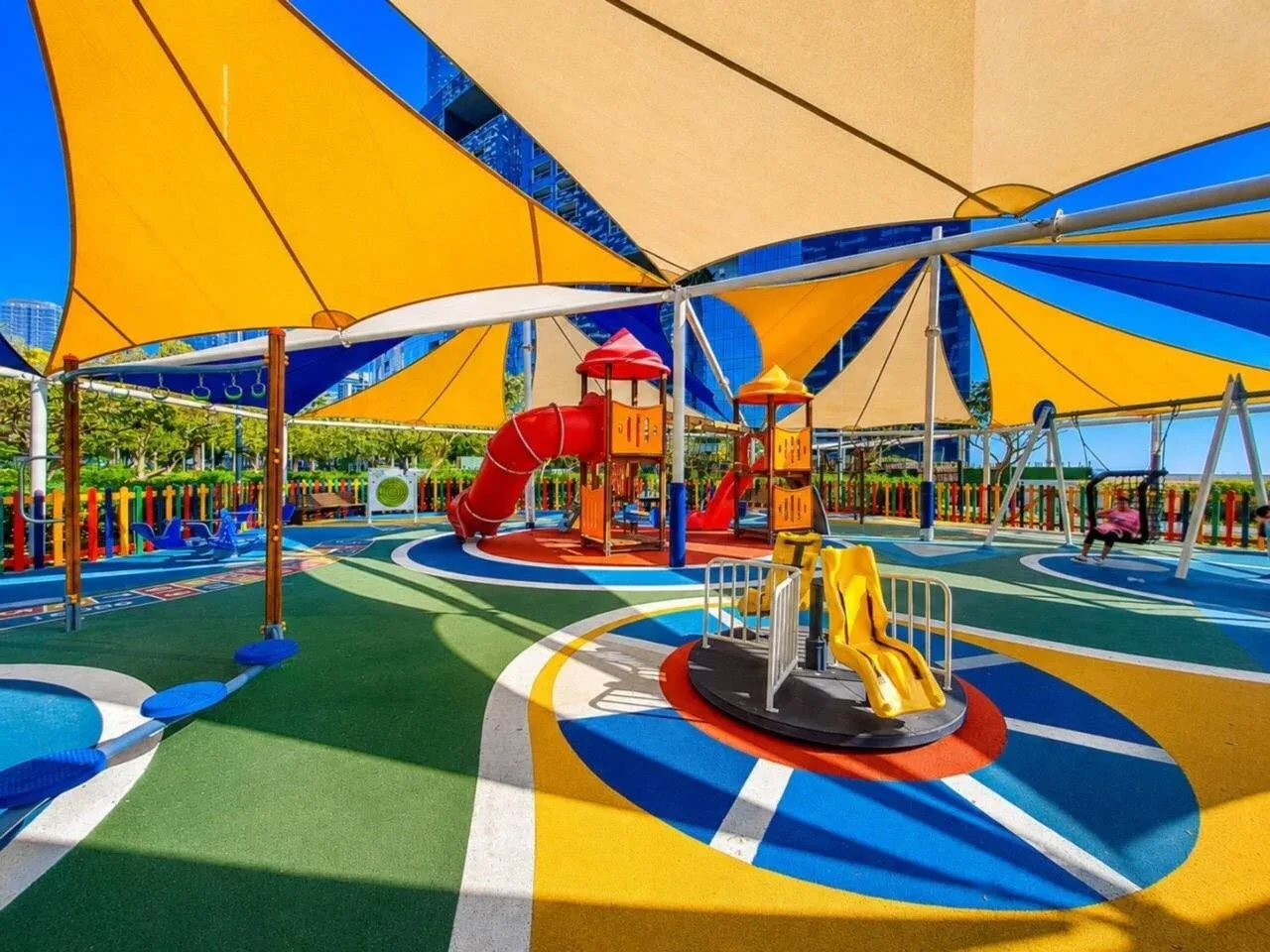 Un parque infantil colorido al aire libre, con toboganes y velas de sombra sobre un suelo estampado.