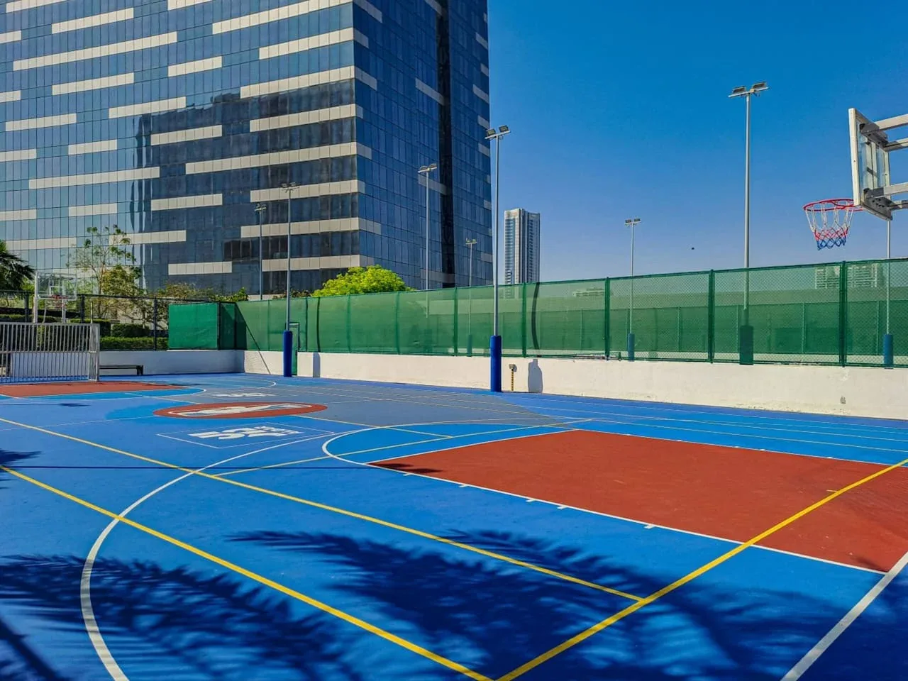 Una cancha de baloncesto al aire libre, con marcas de colores y una canasta junto a un edificio acristalado.