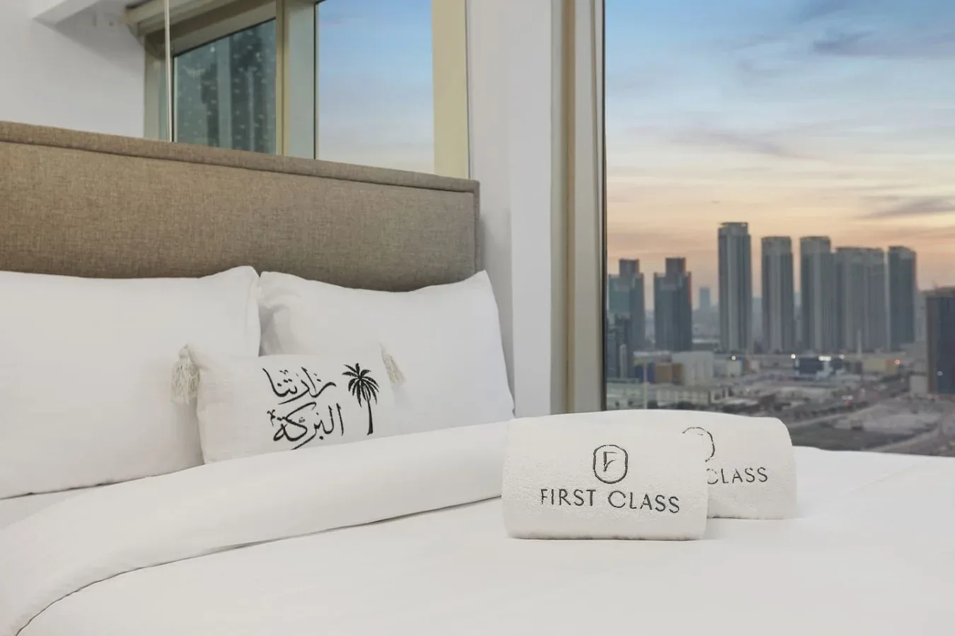 Un dormitorio con ropa de cama blanca junto a una gran ventana con vistas a edificios de la ciudad.