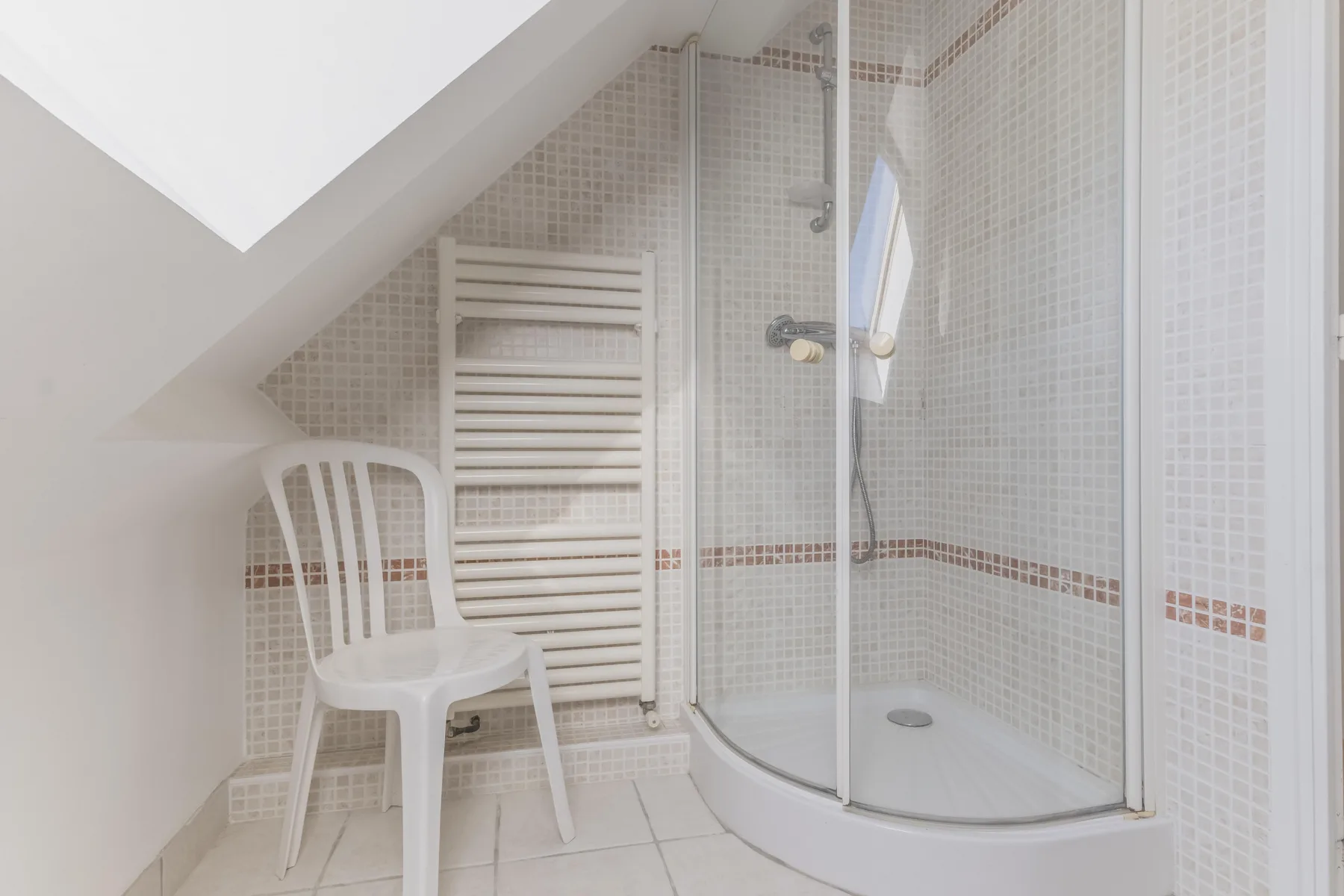 A tiled bathroom with a glass shower and a white chair.