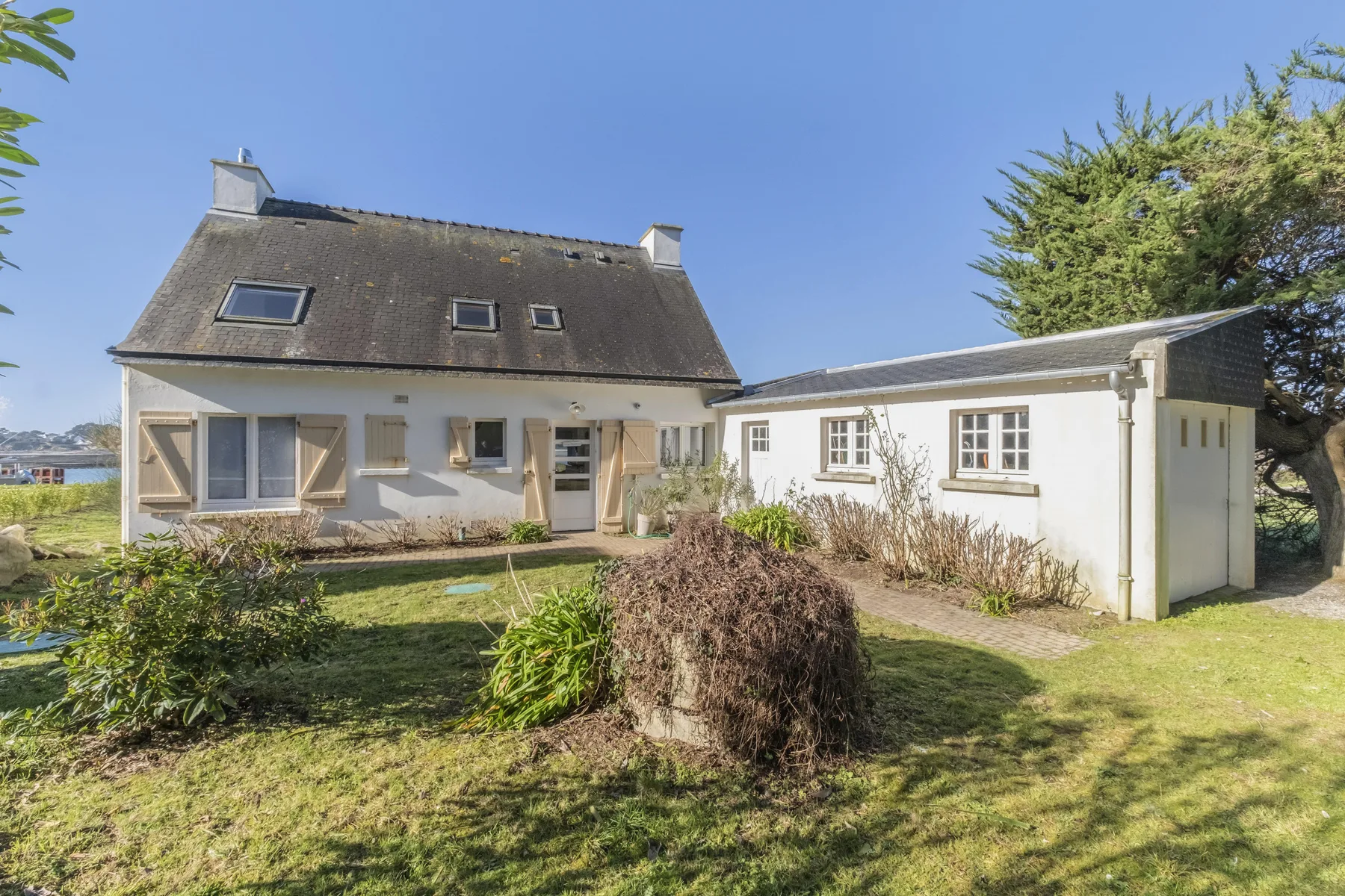 A white house with a dark sloped roof stands beside a sunny garden.