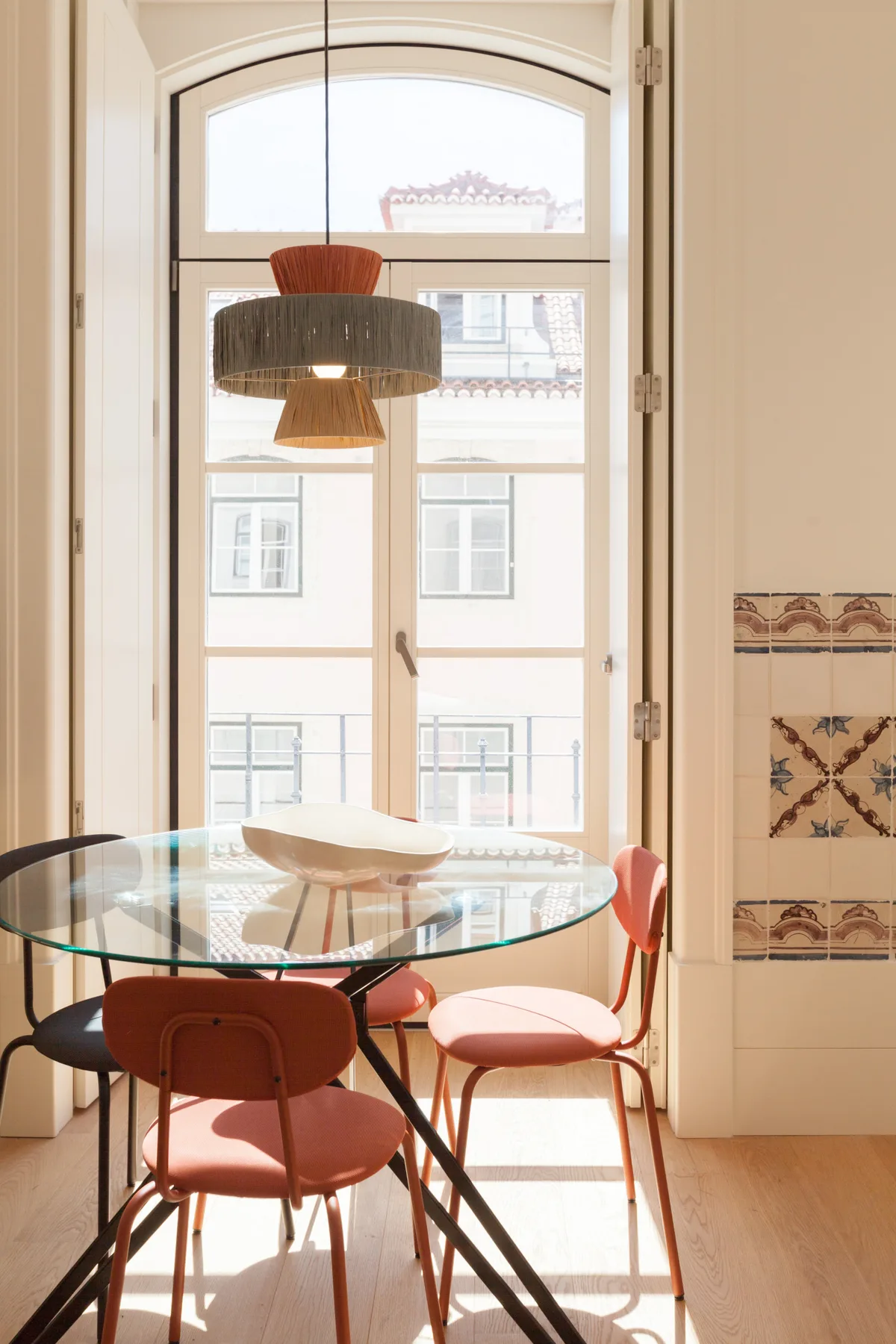 Un comedor con una mesa de vidrio y asientos de colores junto a una ventana alta.