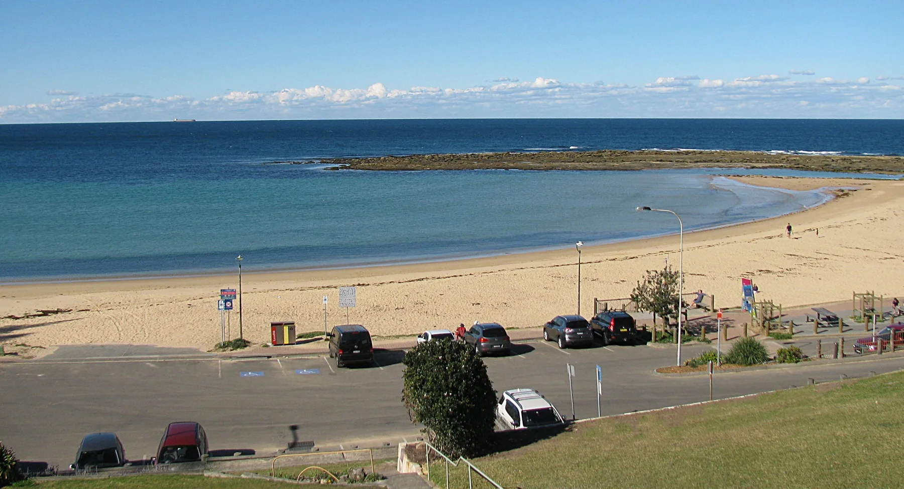 Una playa de arena y aguas azules tranquilas se extienden más allá de una carretera costera.