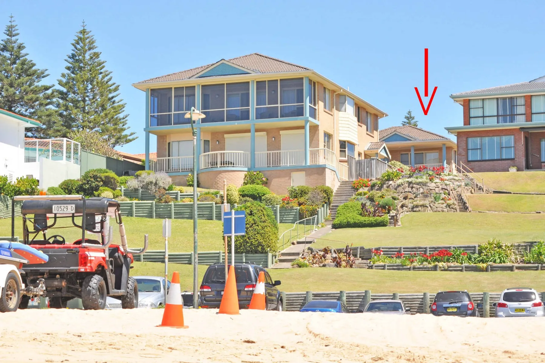 Escena residencial junto a la playa, con casas de varias plantas, jardines en pendiente y vehículos estacionados.