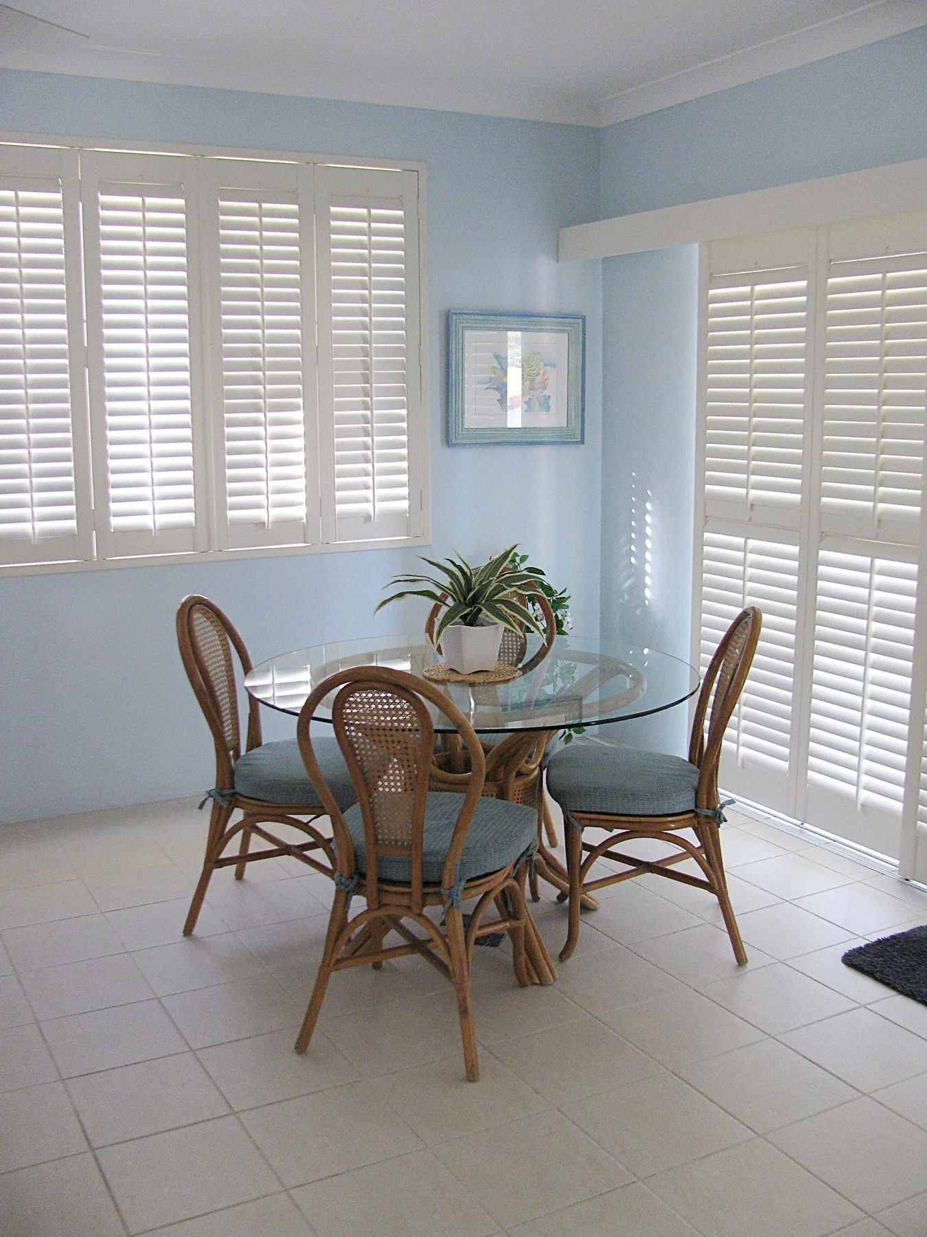 Comedor con mesa de vidrio y asientos de mimbre junto a ventanas con postigos.