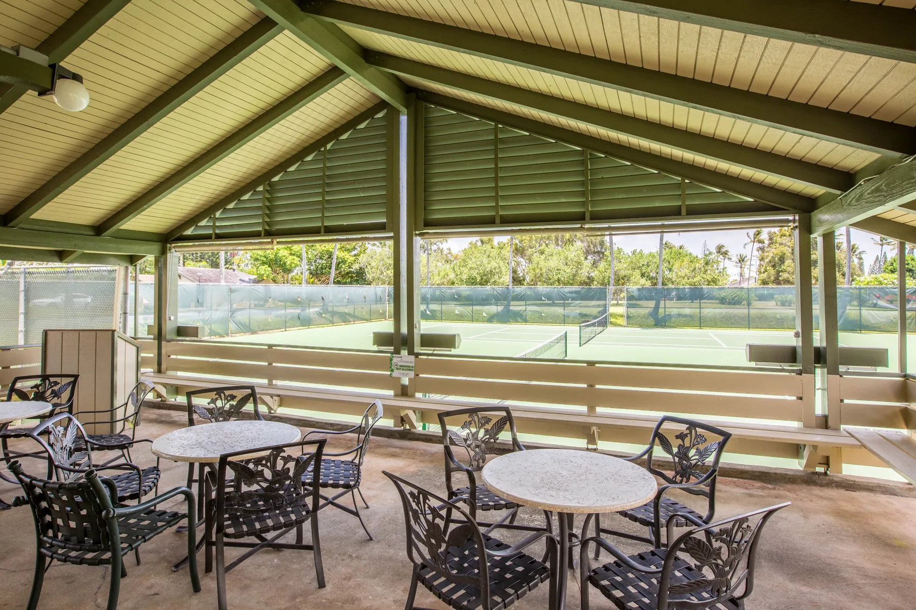 Un patio cubierto con mesas redondas y asientos da a una cancha deportiva cercada.