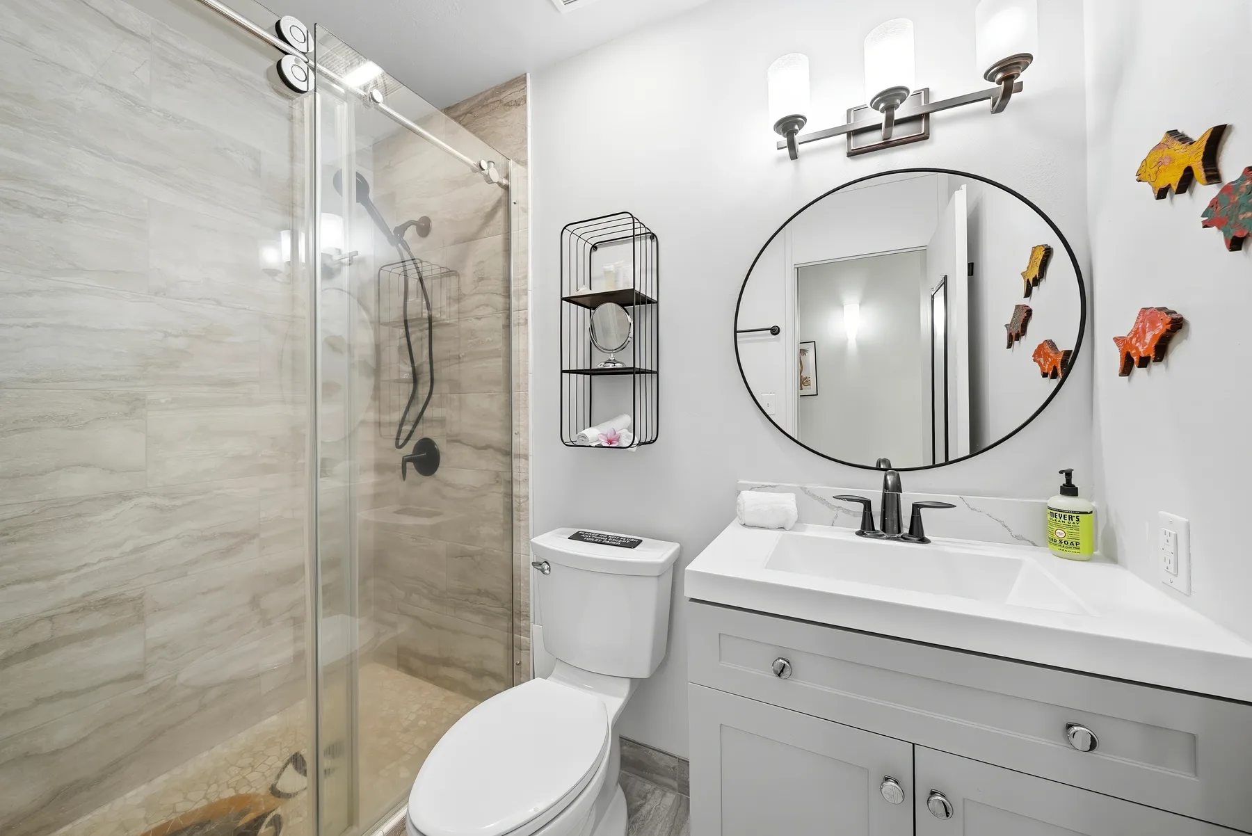 Un baño con ducha de vidrio, inodoro y lavabo bajo un espejo ovalado.