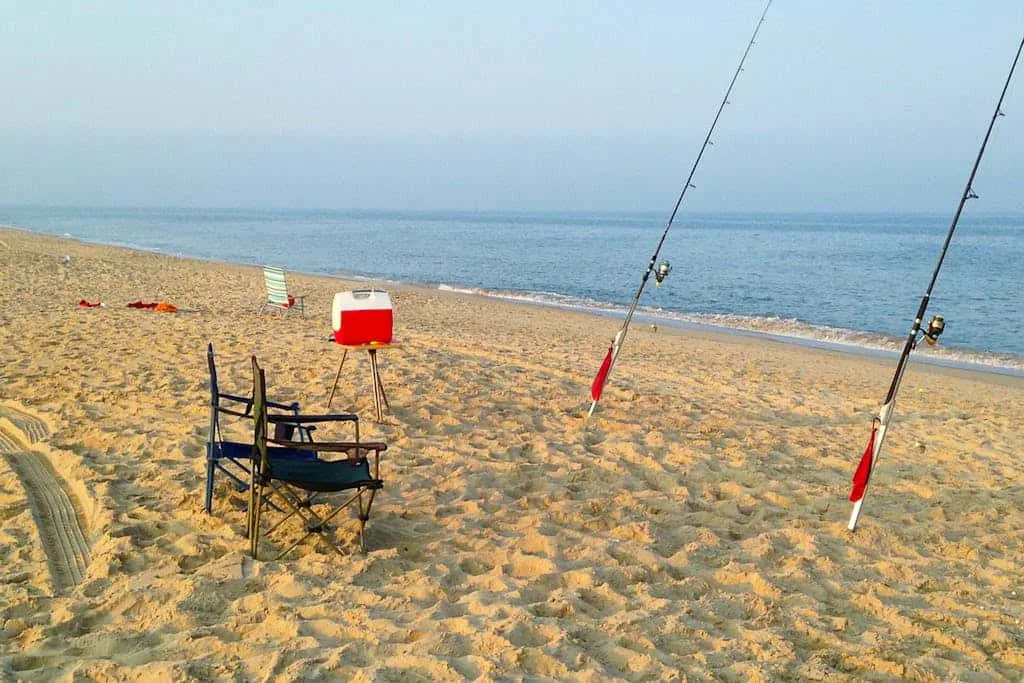 A sandy beach with fishing rods, folding chairs, and a cooler beside the water.