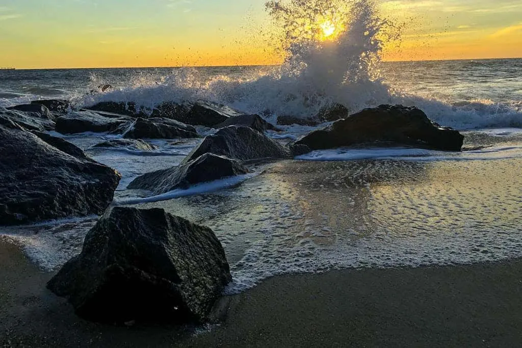 Ocean waves break over dark rocks at sunset.