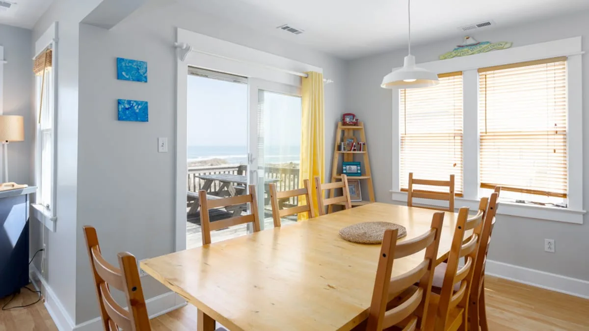 A bright dining area has a large wooden table beside windows and sliding glass doors.