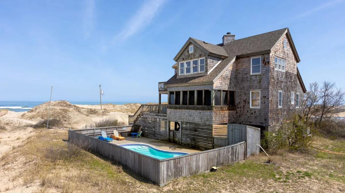 A large shingle-sided house stands beside a fenced pool near sandy dunes.