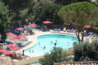 An outdoor swimming pool is surrounded by sun loungers, red umbrellas, and trees.