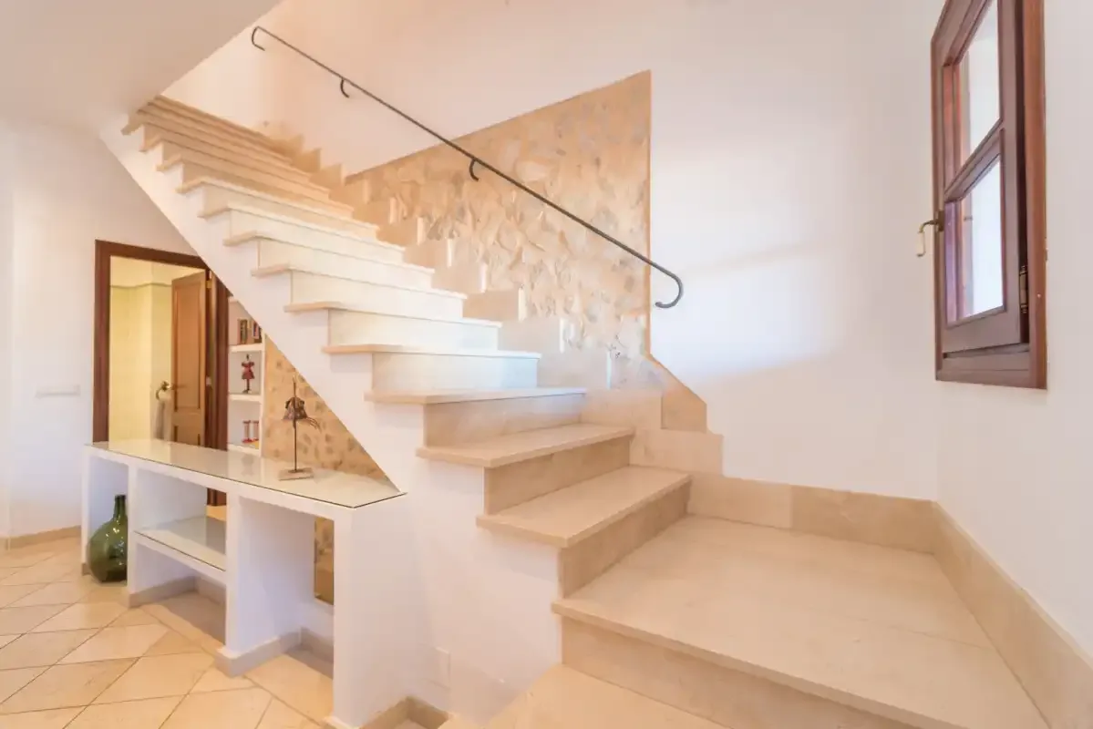 An indoor staircase rises beside a stone-textured wall and a tall window.