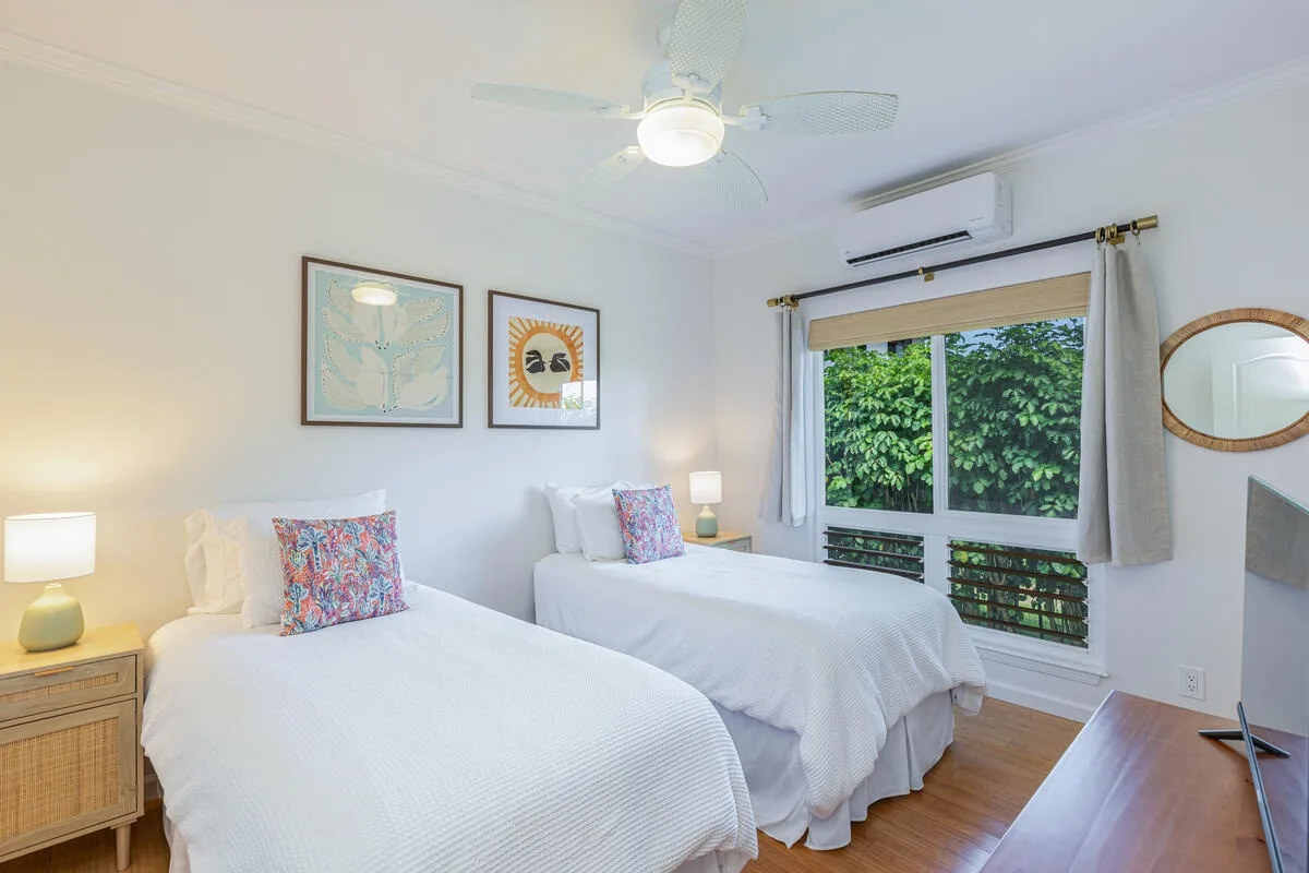 Un dormitorio luminoso con dos camas, cuadros enmarcados y una ventana grande con vegetación al fondo.