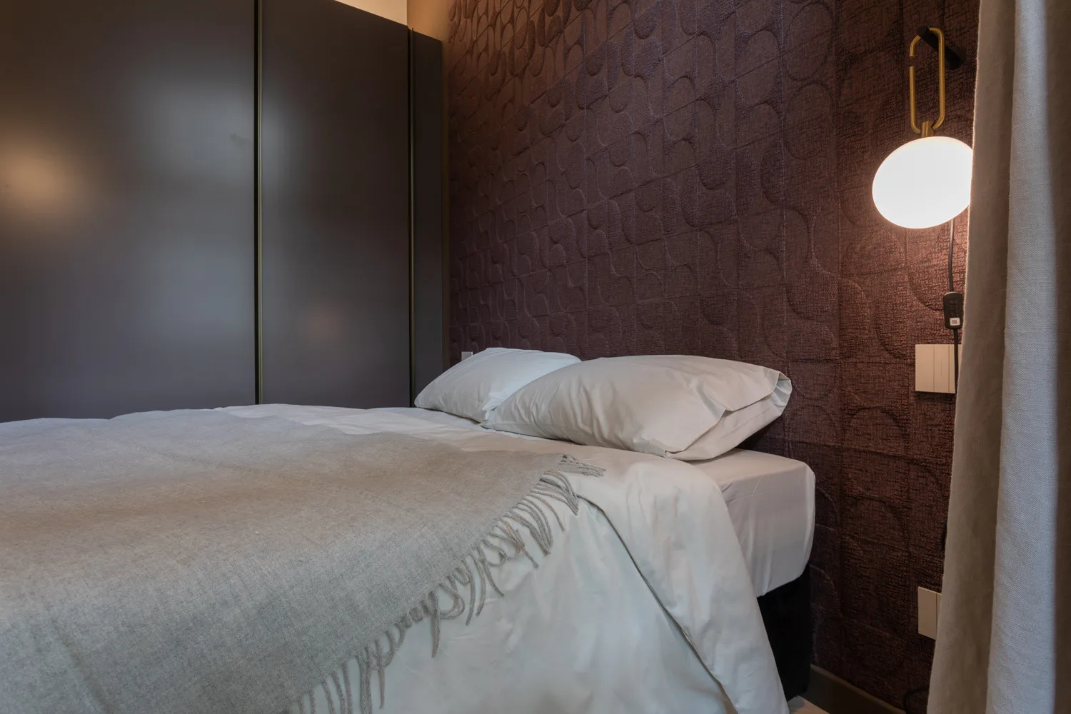 A bedroom with a neatly made bed beside a dark textured wall.