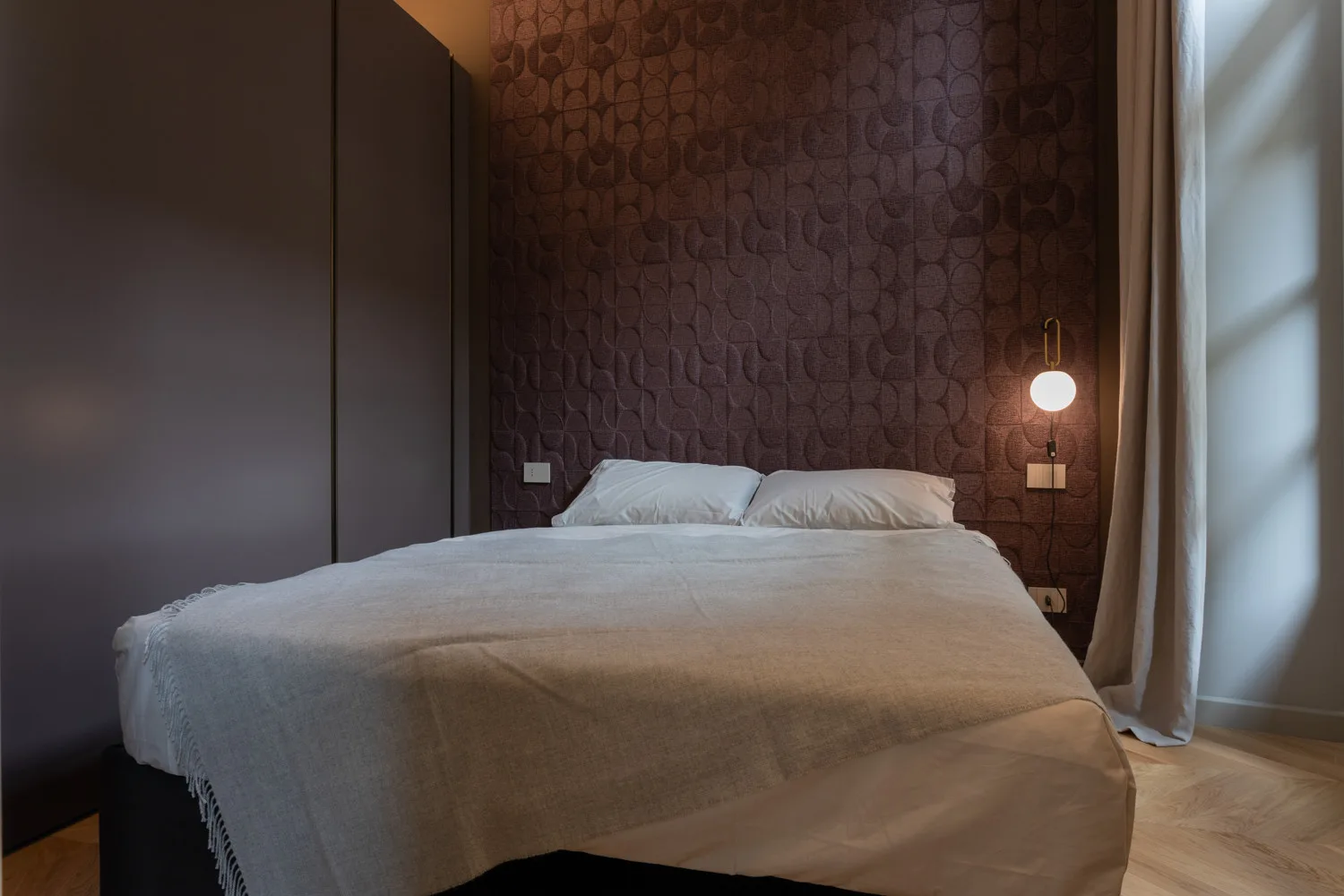 A bedroom with a bed against a patterned wall beside a window.