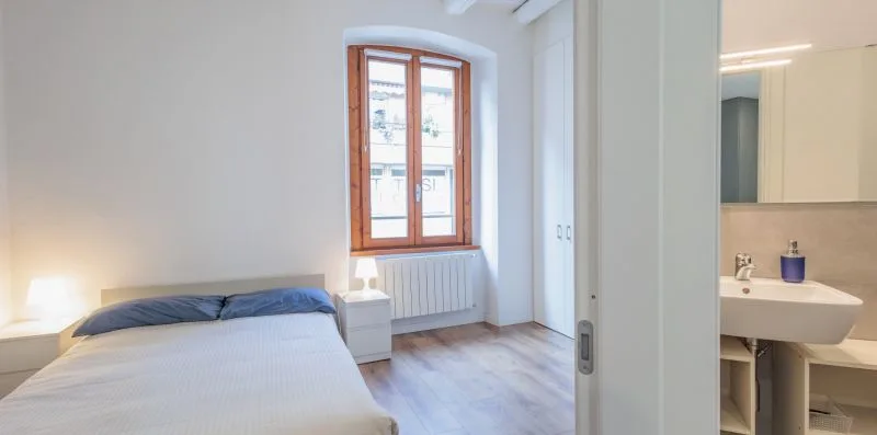 Un dormitorio con una cama junto a una ventana grande y una puerta abierta al baño.