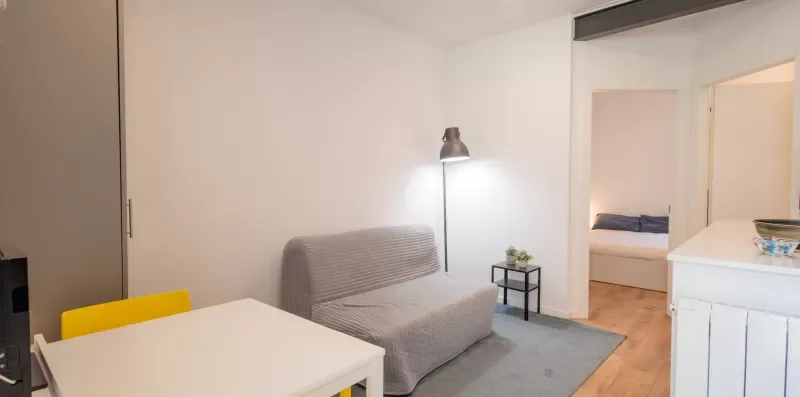 Una pequeña sala de estar con asientos, una mesa y una puerta abierta hacia un dormitorio.