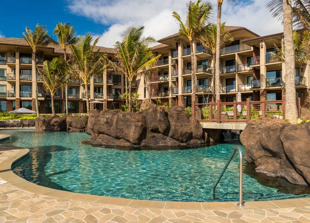 Un edificio de resort se alza detrás de una piscina rodeada de rocas y palmeras.