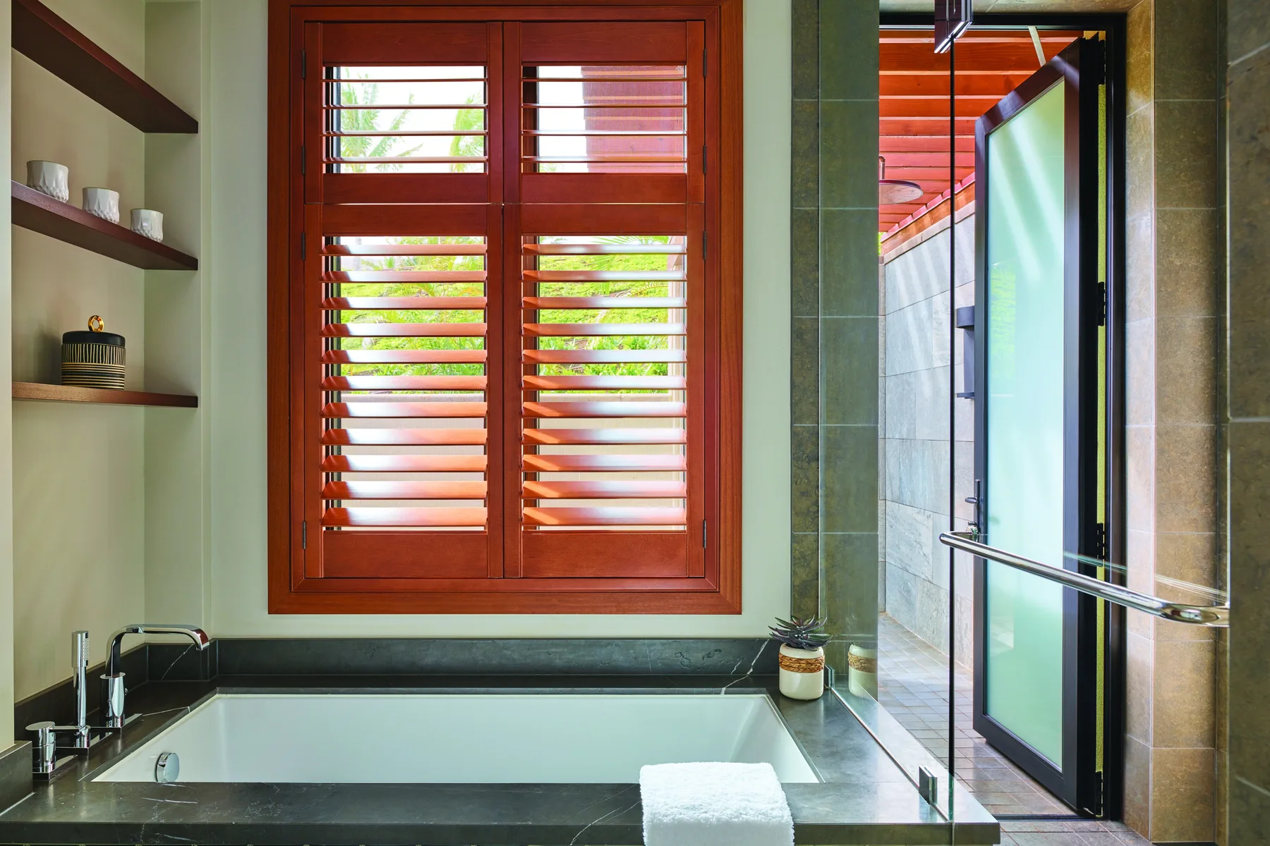 Un baño con bañera bajo una ventana de lamas de madera y una zona de ducha abierta.