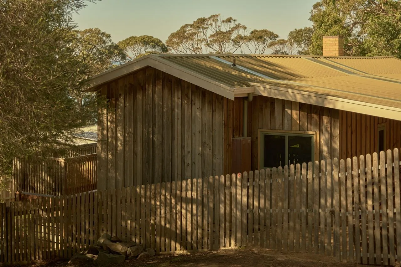 A wooden house with a sloping roof stands behind a picket fence.