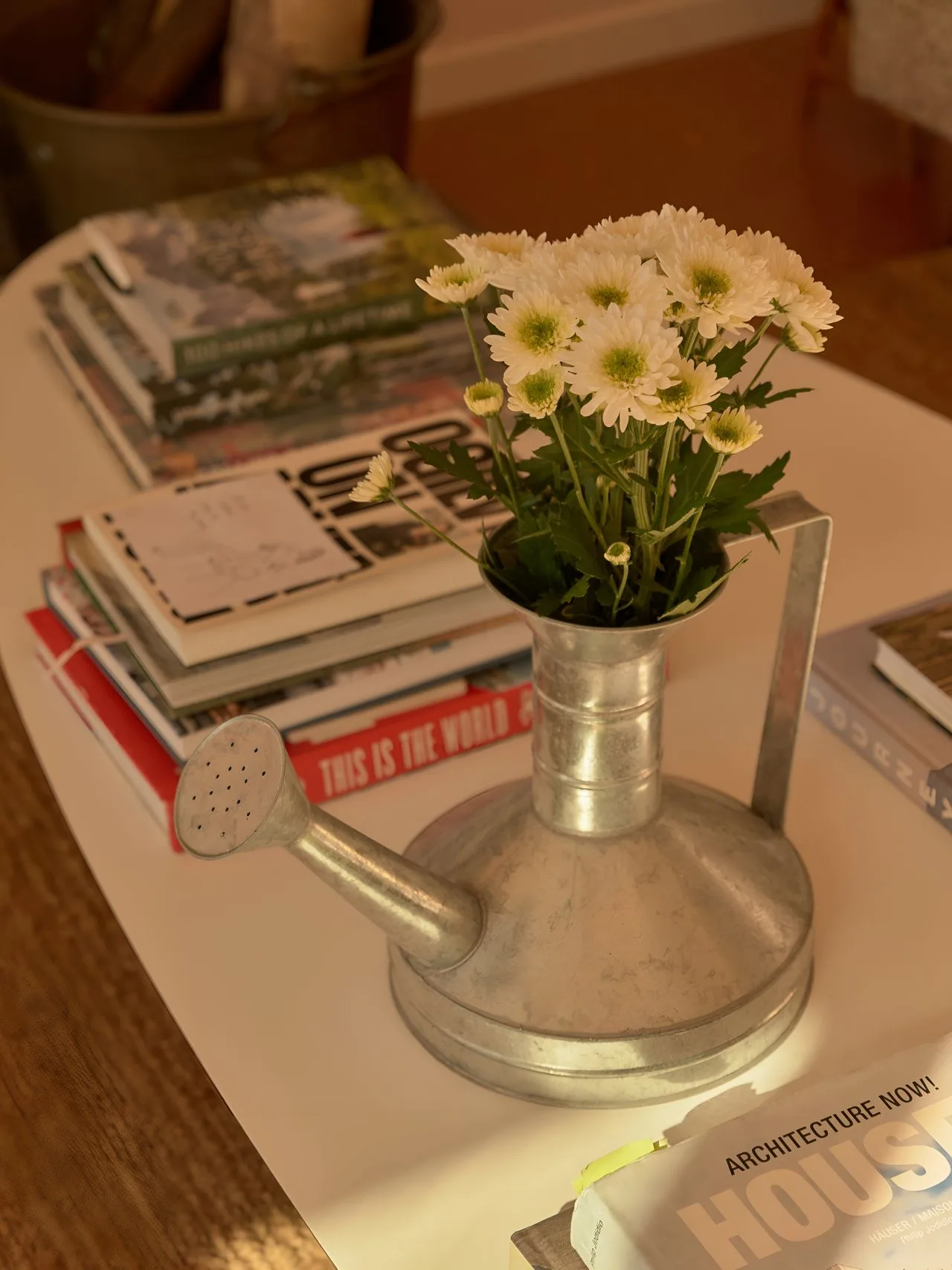 A flower arrangement sits in a metal watering can on a table beside magazines.