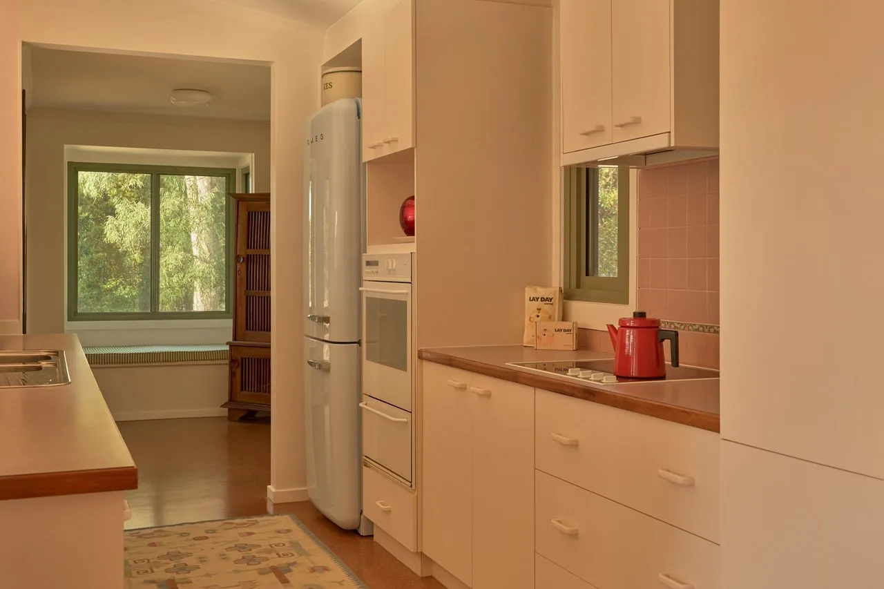 A kitchen with white cabinets and a refrigerator, opening onto a room with large windows.