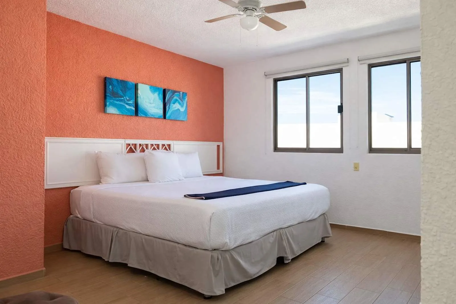 A bedroom with a bed beneath colorful wall art and two windows.