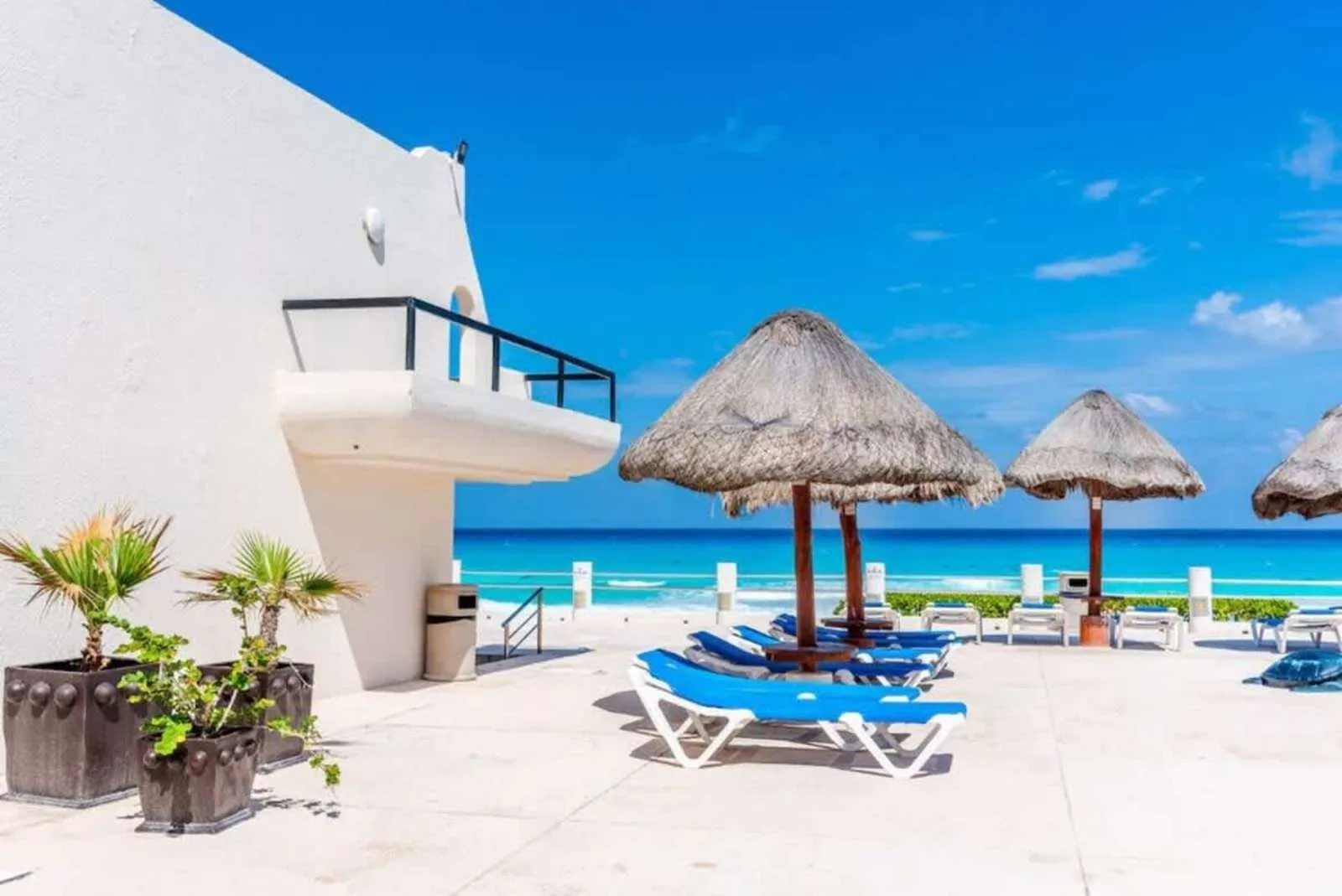 A sunny seaside pool deck has lounge seating beneath thatched parasols.