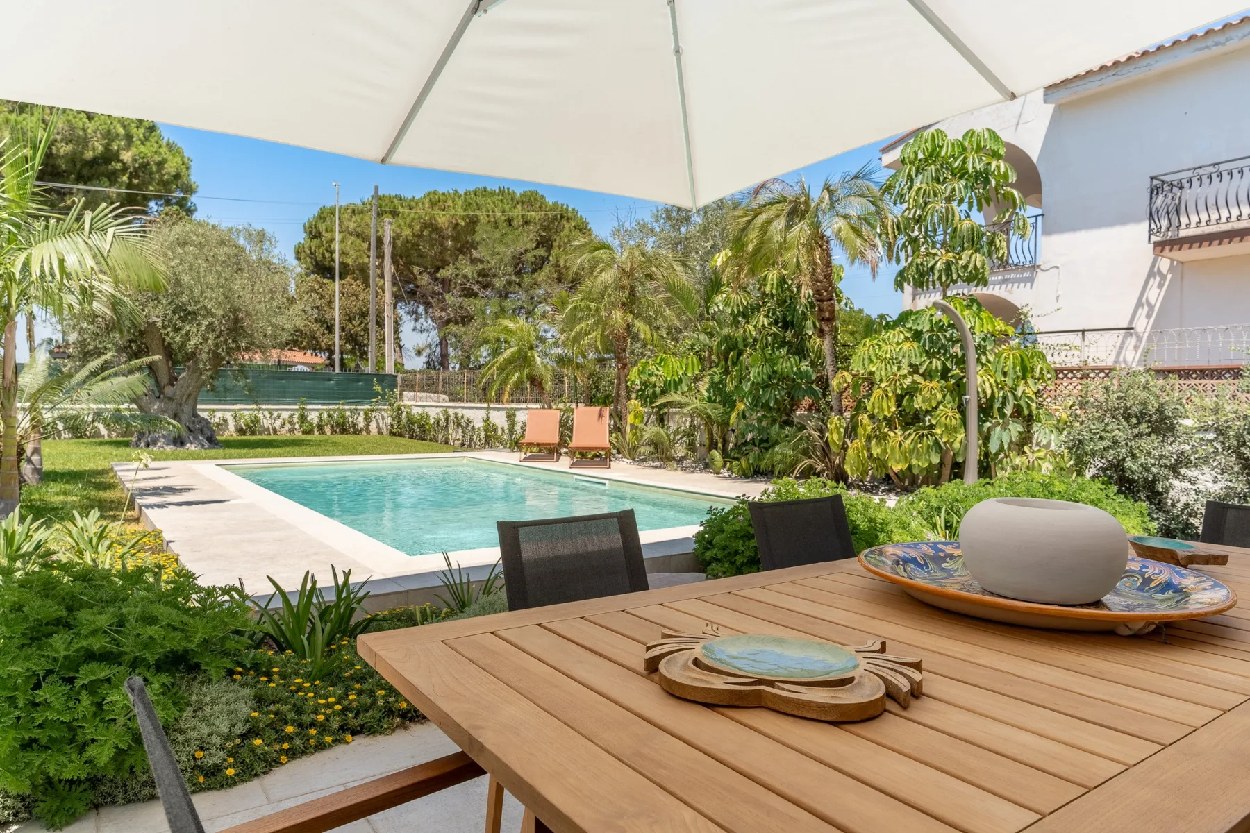 A garden patio overlooks a swimming pool, with a dining table in the foreground.