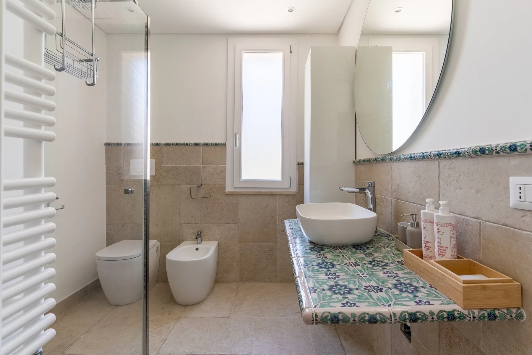 A bright bathroom with a glass shower, wall-mounted sink, and window.