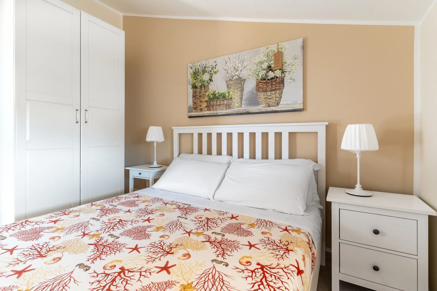 A bedroom with a bed, white bedside tables, and floral wall art.