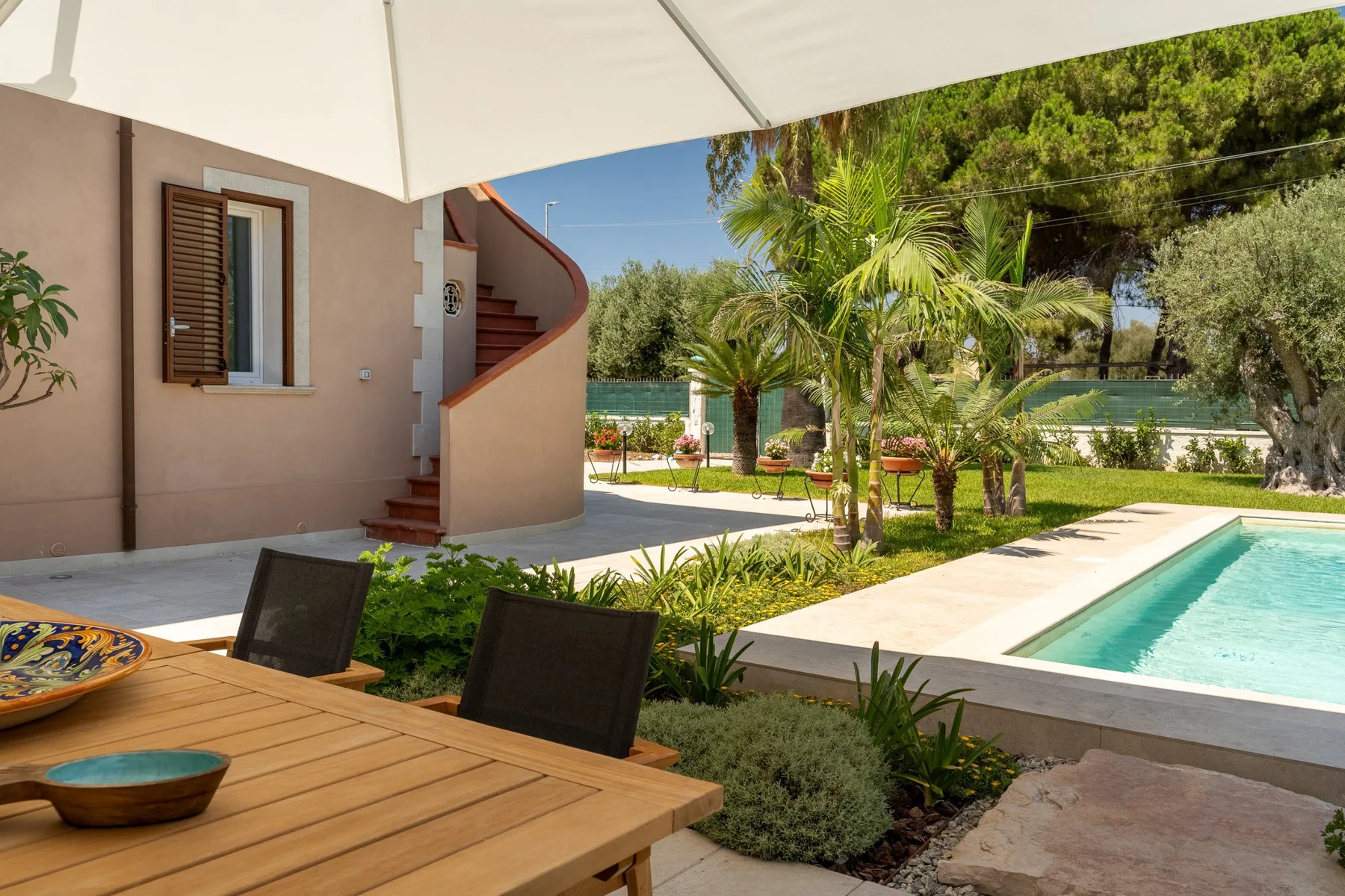 A sunny outdoor patio with a wooden table, pool, and palm trees.
