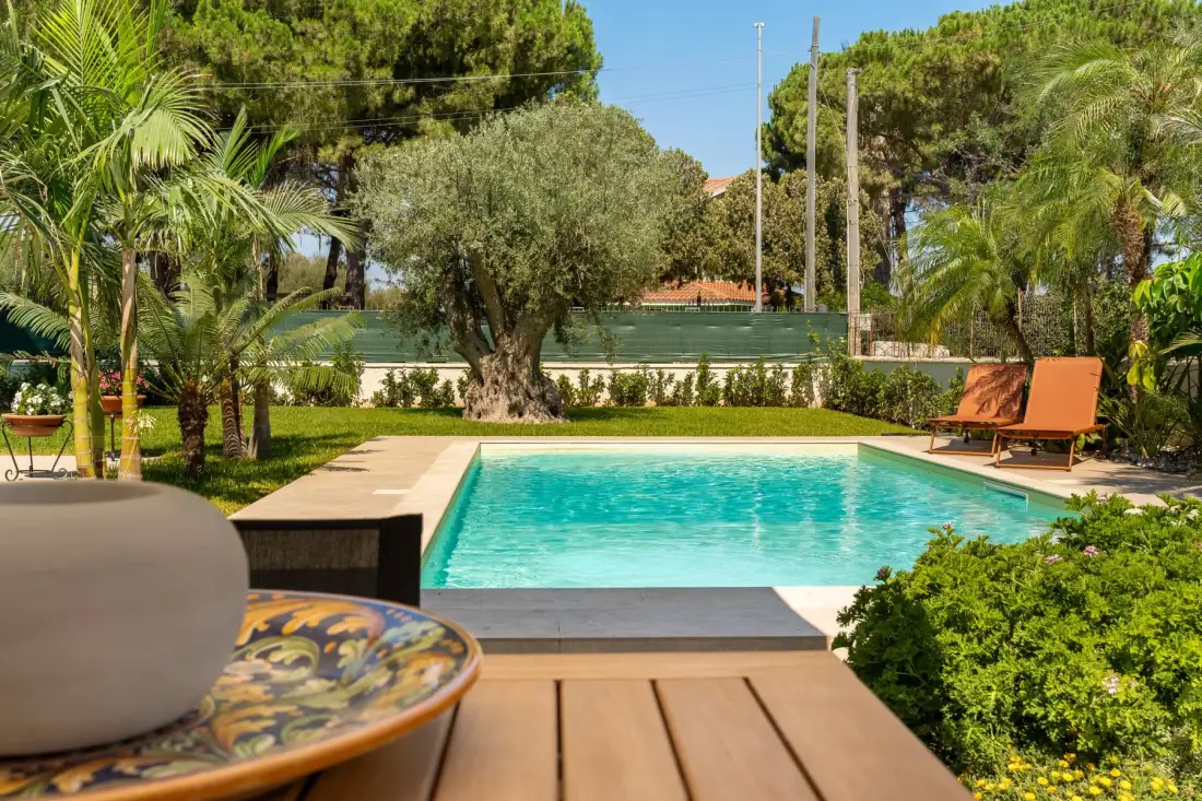 An outdoor swimming pool sits among trees, shrubs, and patio seating.