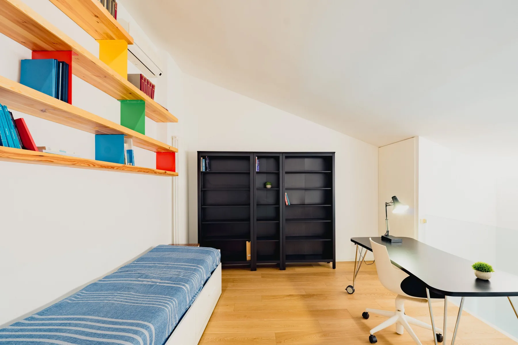Un estudio luminoso con una cama individual, escritorio y estantes en la pared.