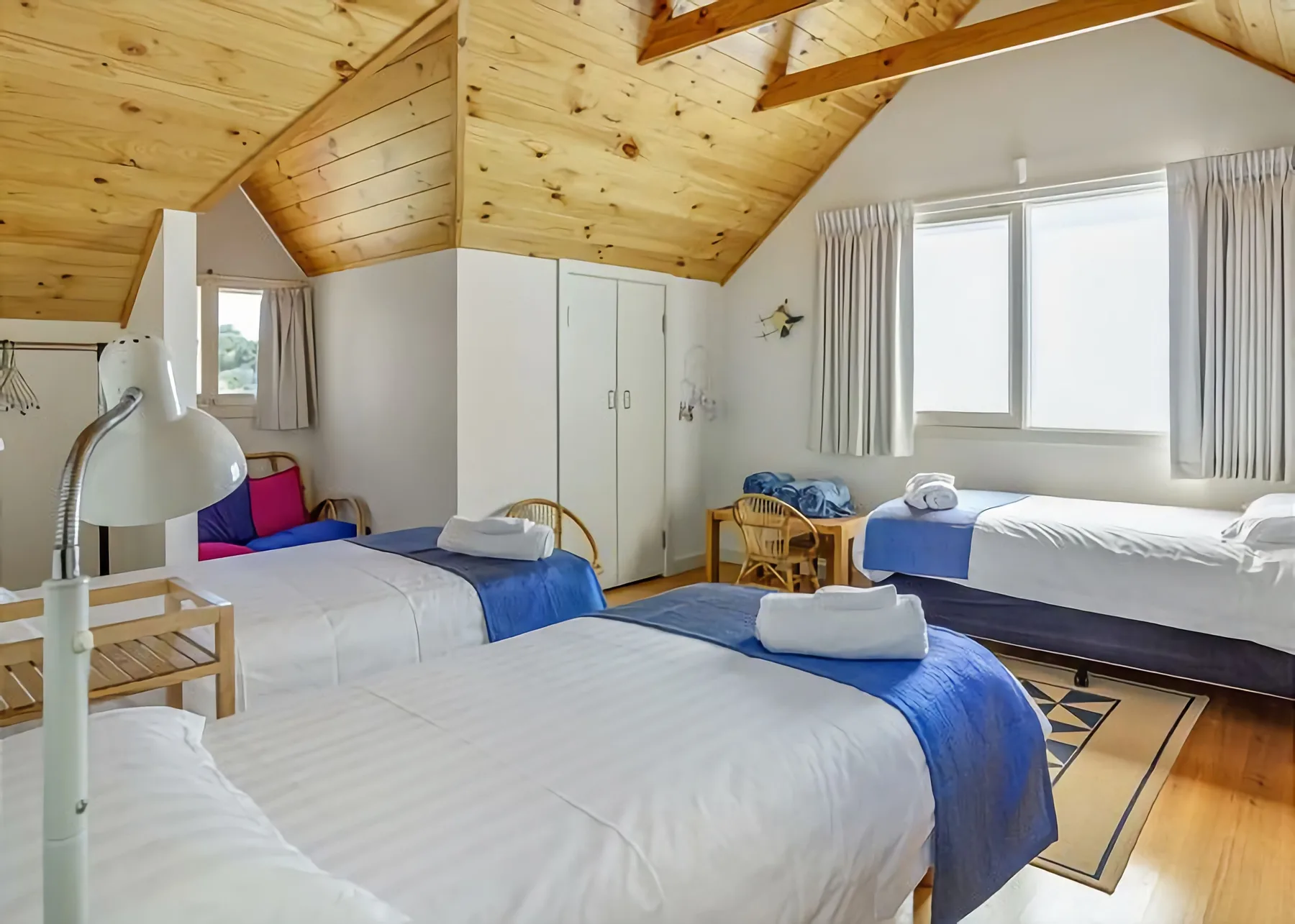 Un dormitorio luminoso con varias camas, cubrecamas azules y un techo de madera inclinado.
