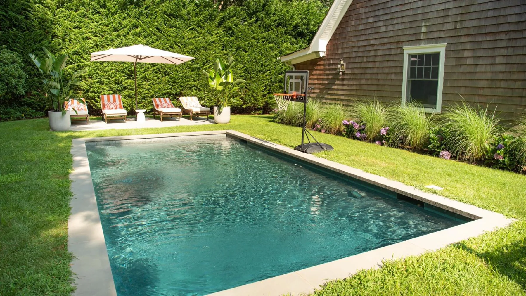 Una piscina al aire libre está junto a un jardín y un edificio con revestimiento de tejas.