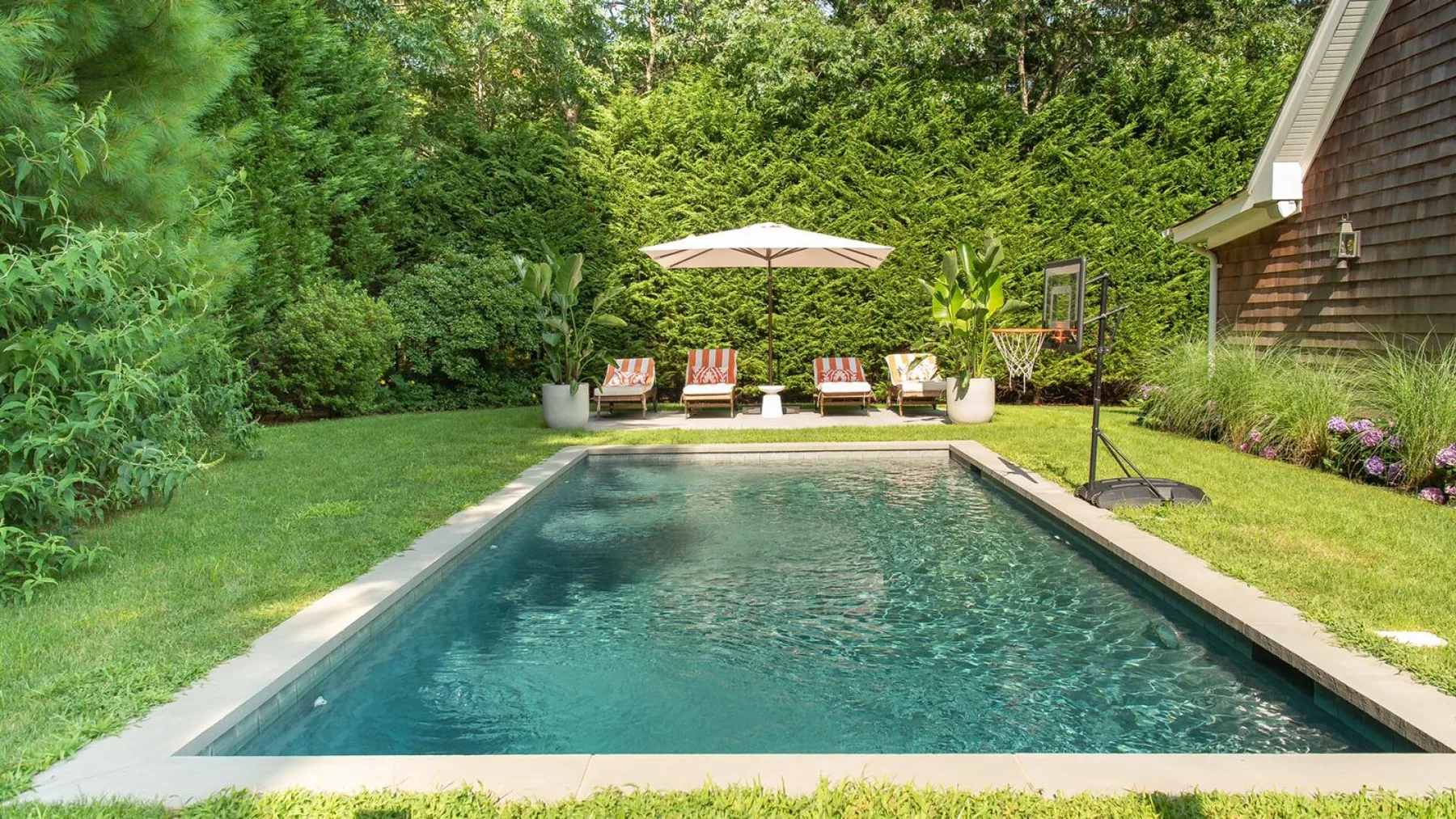 Una piscina al aire libre está rodeada de césped, árboles y asientos de patio.