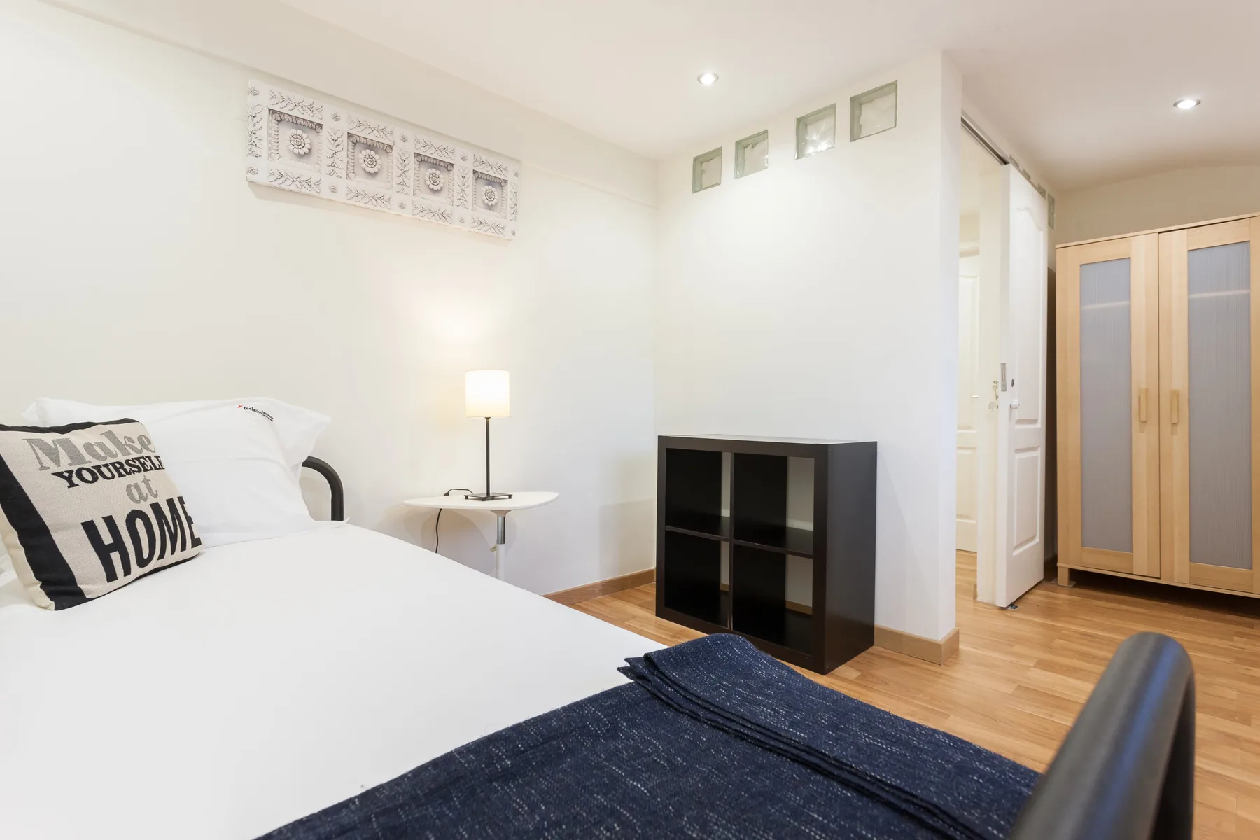 Un dormitorio luminoso con una cama, una mesita y una estantería oscura.
