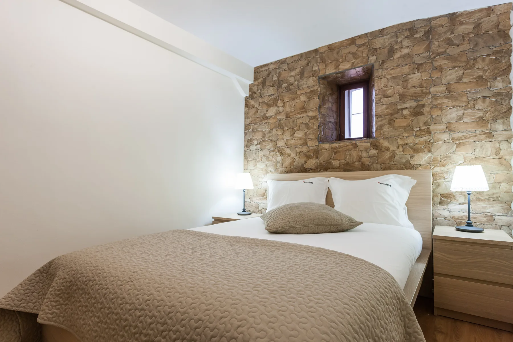 Un dormitorio con una cama junto a una pared de piedra y una ventana pequeña.