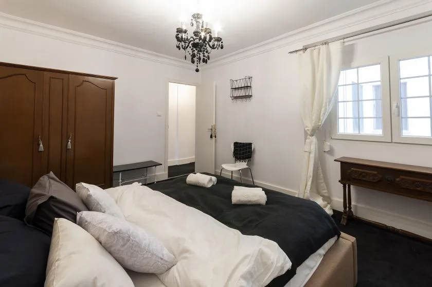 A bedroom with a large bed, dark bedding, and windows with light curtains.