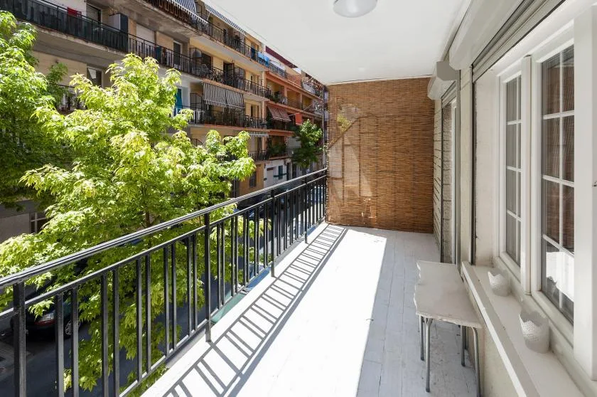 A sunlit balcony with a black railing, neighboring buildings, and leafy trees.