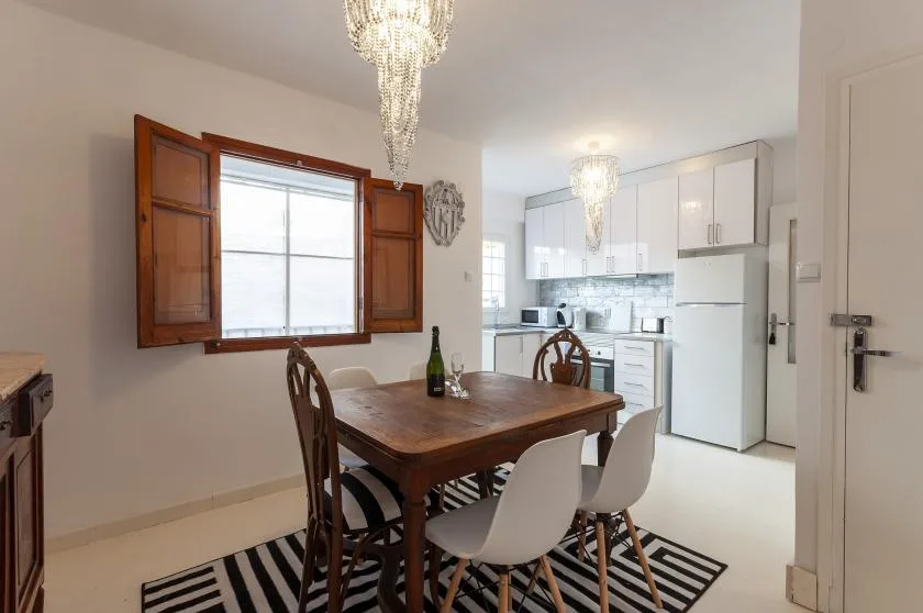 A dining area opens onto a bright kitchen, with a wooden table beneath a chandelier.