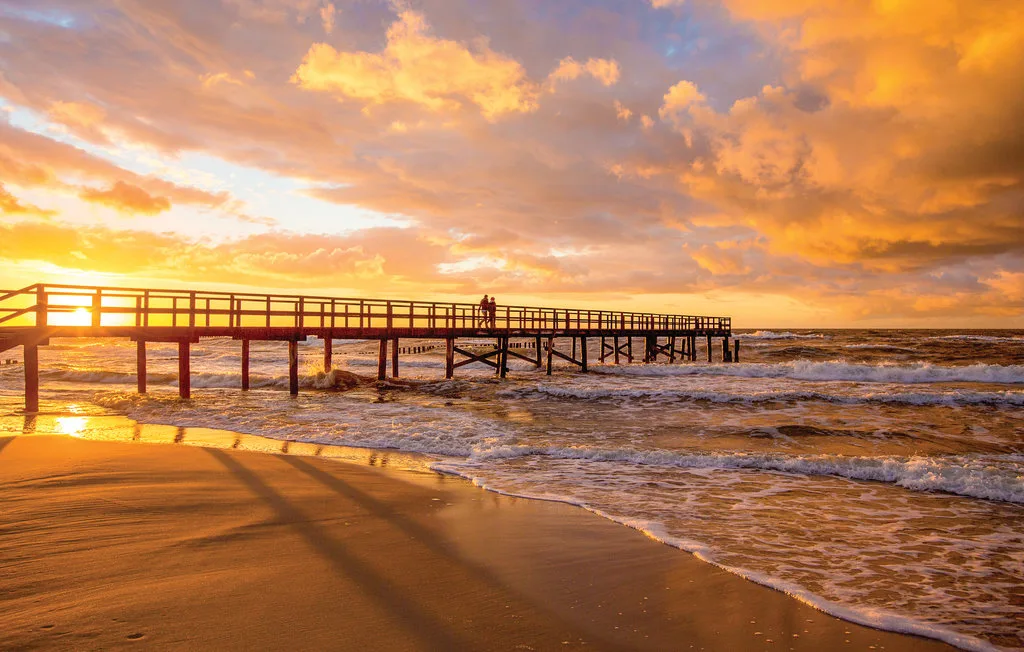 A wooden pier extends over the beach beneath a colorful sunset sky.