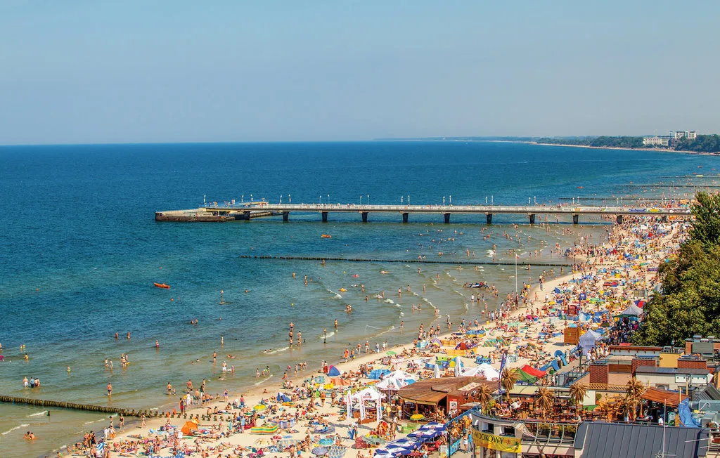 A crowded beach stretches along the blue sea, with a pier extending offshore.