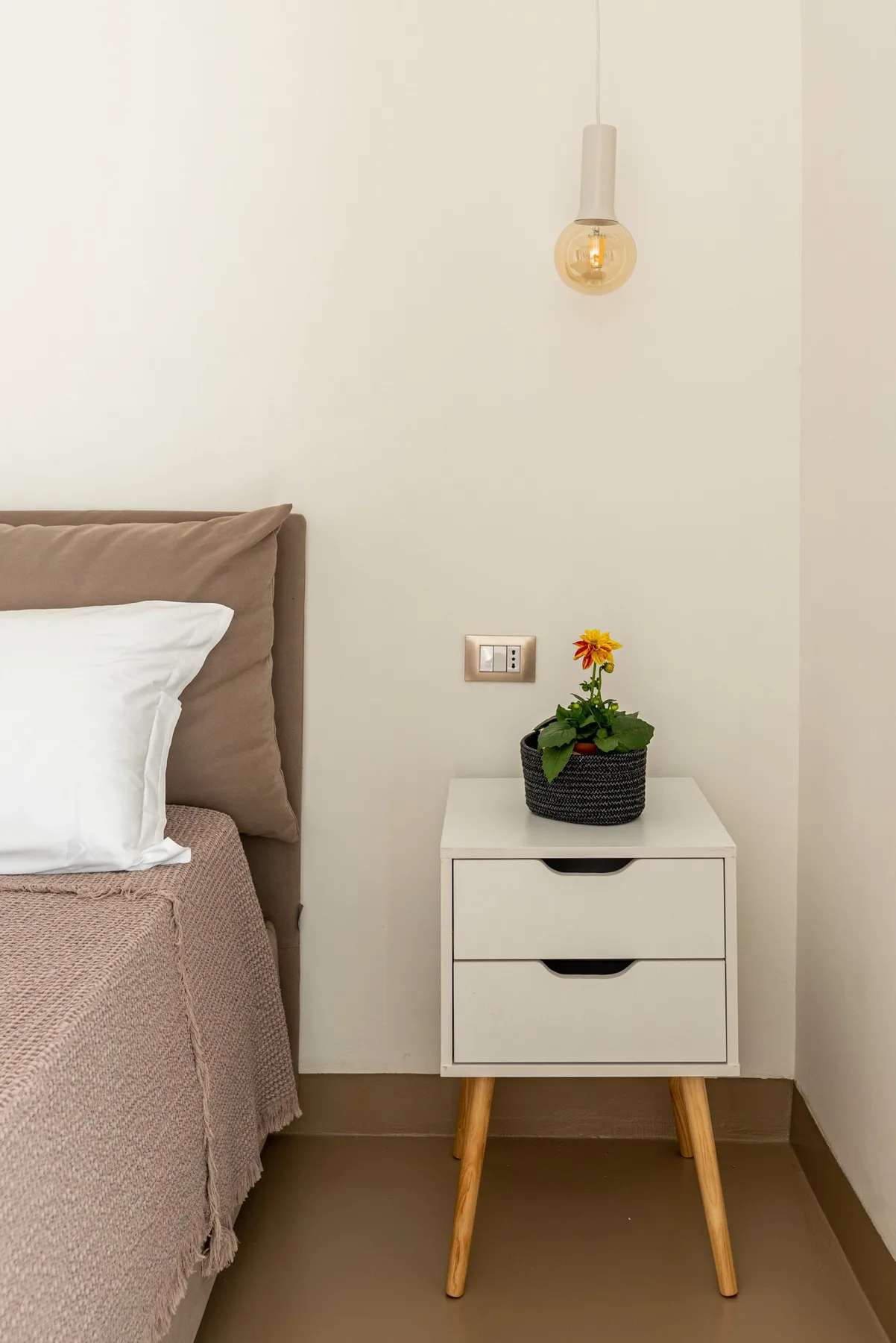 Rincón de dormitorio con una cama junto a una mesita de noche y una flor en maceta.