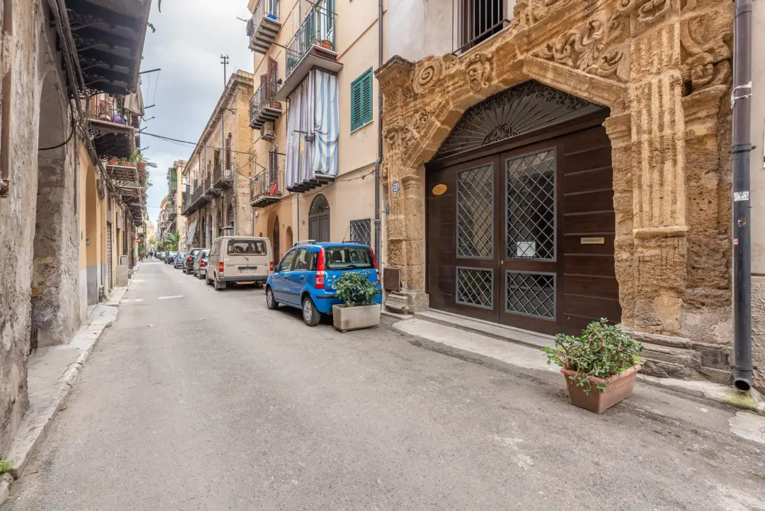 A narrow street lined with buildings, parked cars, and an ornate stone doorway.